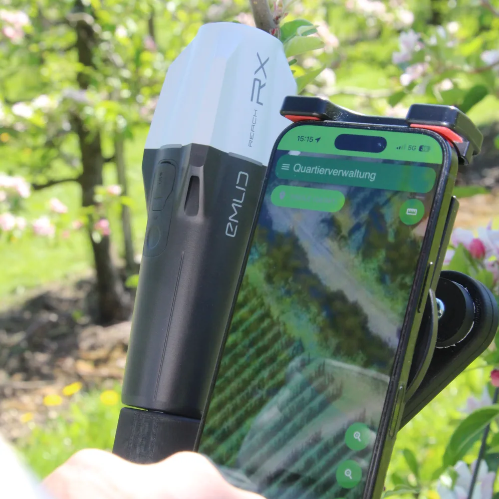 Eine Hand hält den DINGO im blühenden Obstbau: RTK-Antenne mit angesetztem Smartphone, auf dem Display die Quartierverwaltung mit der Anlage in der Karte.